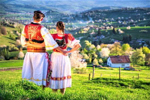 pár v kroji stojaci na lúke a pozerajúci sa na slovenskú dedinu